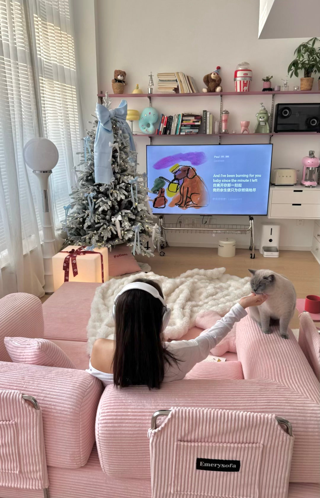 Woman sitting on a pink couch with a cat, watching TV in a living room decorated for Christmas.